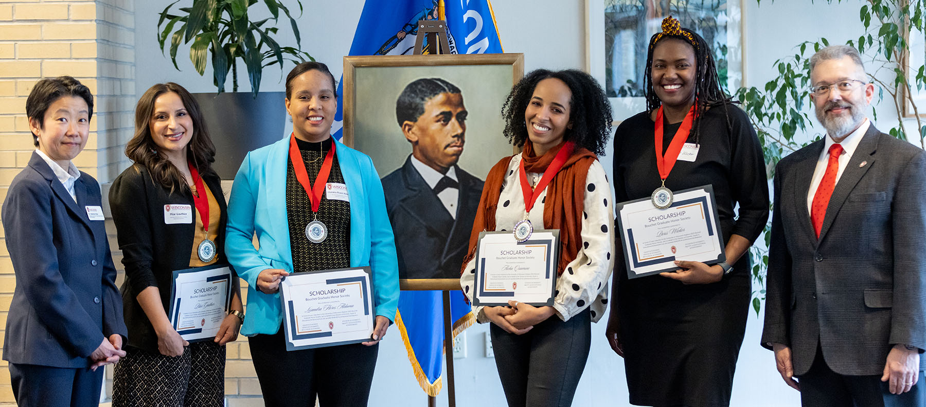 Six people pose for a photo next to a framed, painted portrait. Four of the people are wearing medals and holding certificates.