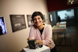 Gabriela Yepes-Rossel stands in a gallery space and leans forward on a pedestal displaying what appears to be an old camera. She is smiling.